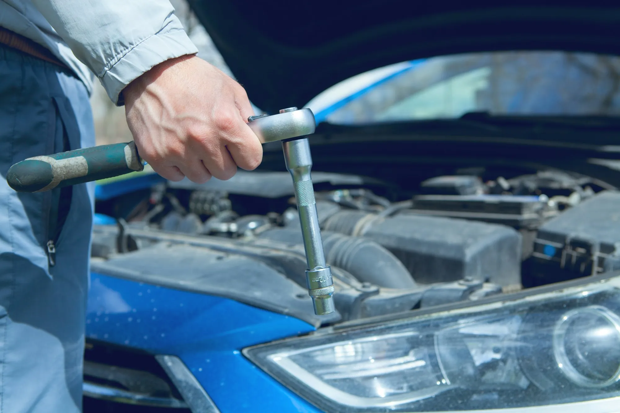 Hand turning a ratchet on an engine next to a blue car fender